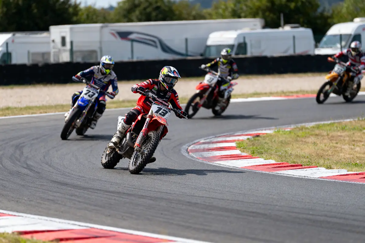 Motards en course sur circuit, virage serré, supemoto, asphalte, journée ensoleillée, arrière-plan avec camionnettes blanches.