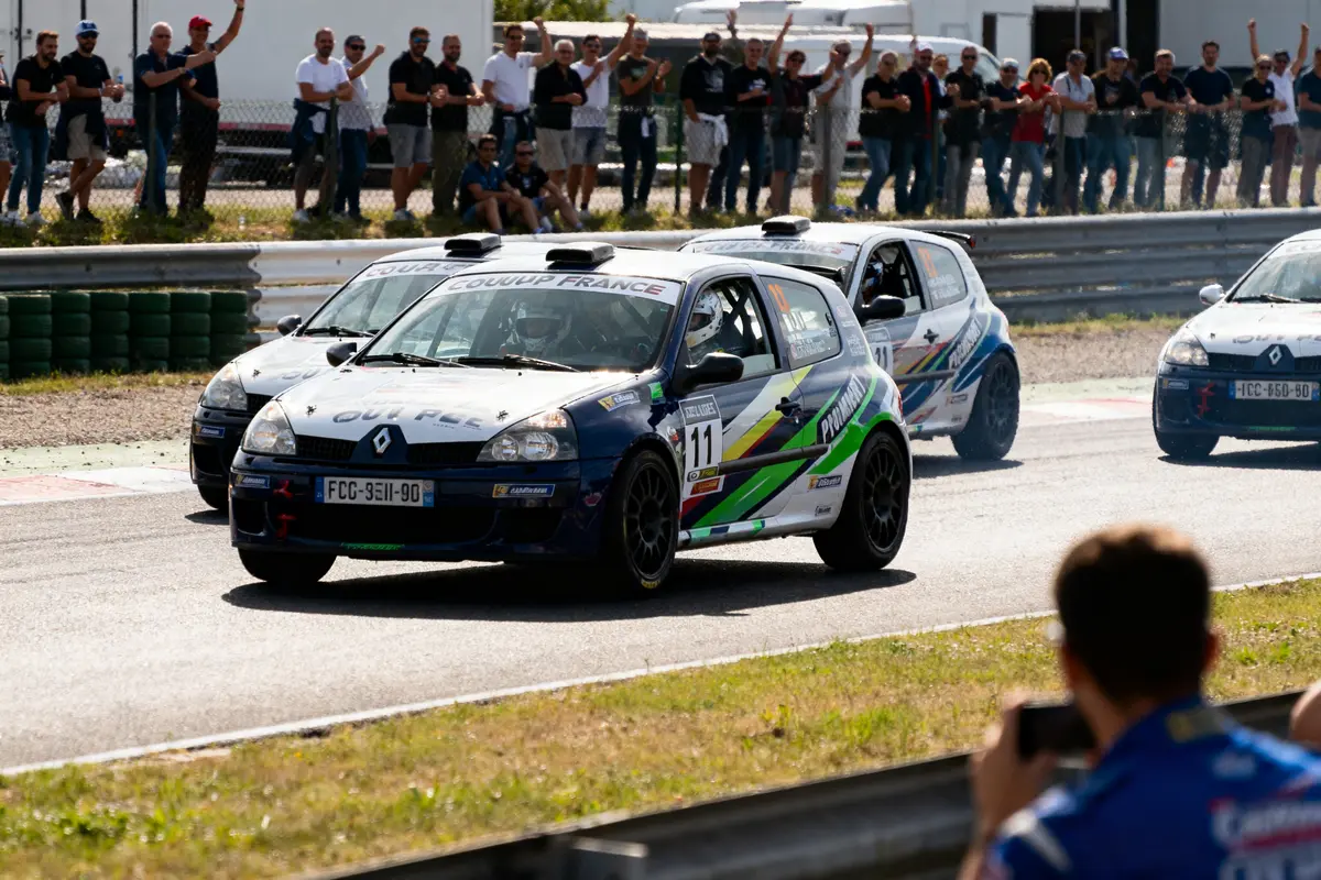Voitures de course Renault Clio sur circuit, foule en arrière-plan, spectateur prenant des photos au premier plan.