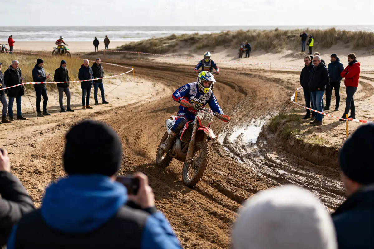 Course de motocross sur sable, plusieurs pilotes en action, spectateurs observant, plage en arrière-plan, ciel nuageux.