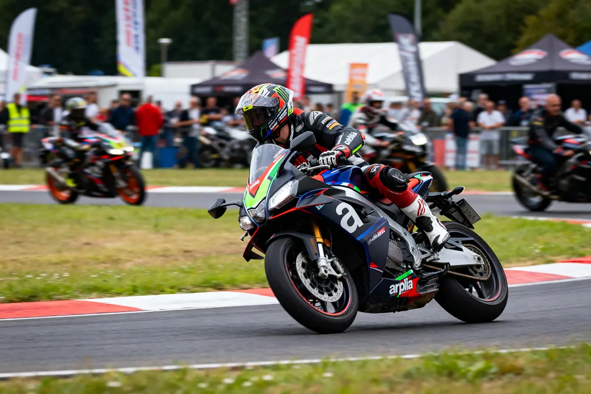 Motard en compétition sur circuit, moto Aprilia, virage, spectateurs flous en arrière-plan, ambiance de course dynamique.