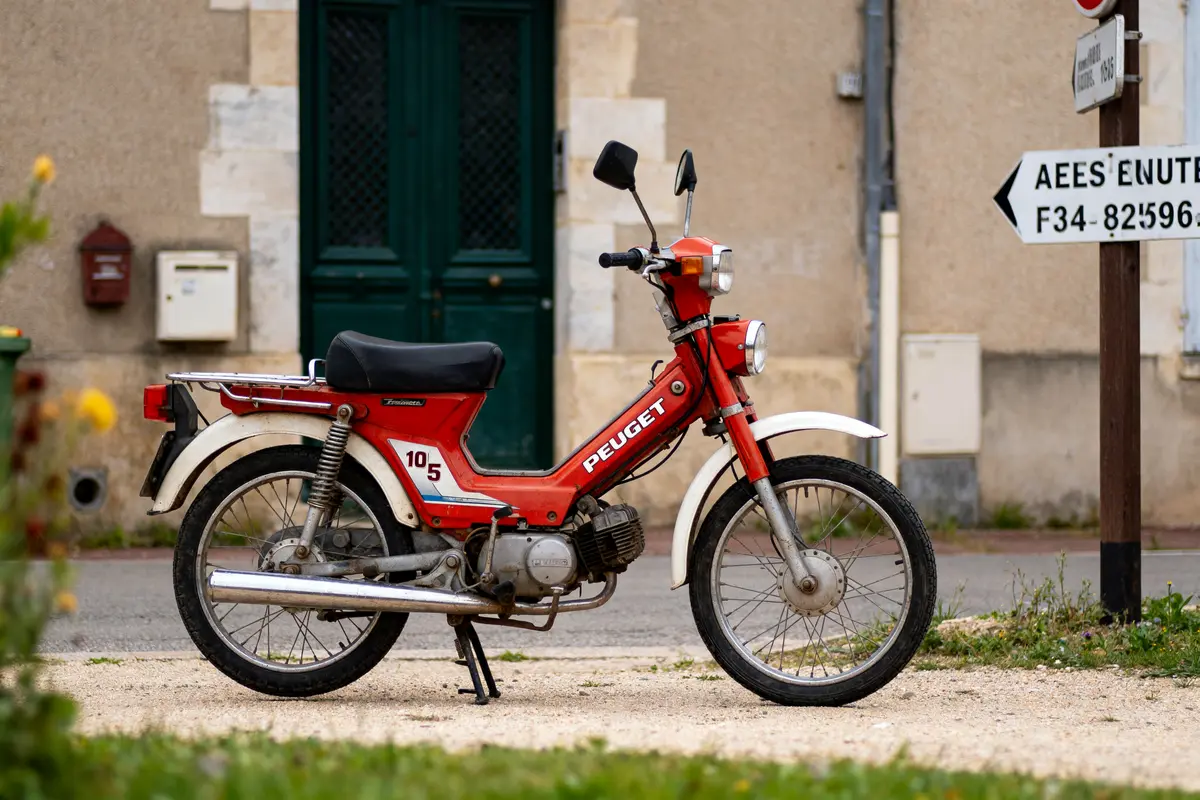 Mob d'époque rouge Peugeot stationnée devant bâtiment beige, panneau indicateur à droite.