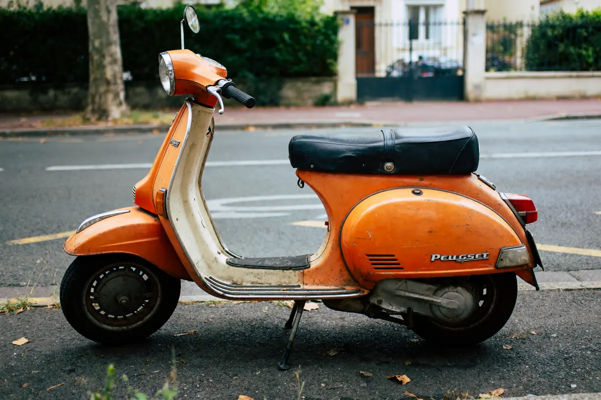 Scooter orange vintage stationné sur le trottoir devant une rue, entouré de feuillage et clôtures.