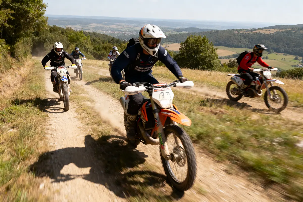 Motocyclistes tout-terrain en course sur un chemin de terre entouré de verdure et de collines.