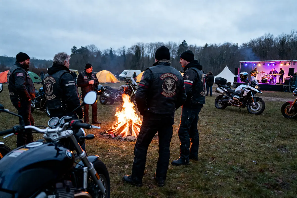 Groupe de motards autour d'un feu de camp, tentes et concert en arrière-plan, ambiance rassemblement extérieur.