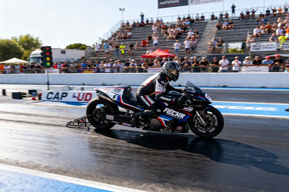 Motard en combinaison noire sur une moto de course, piste de drag, public en tribunes, jour ensoleillé.
