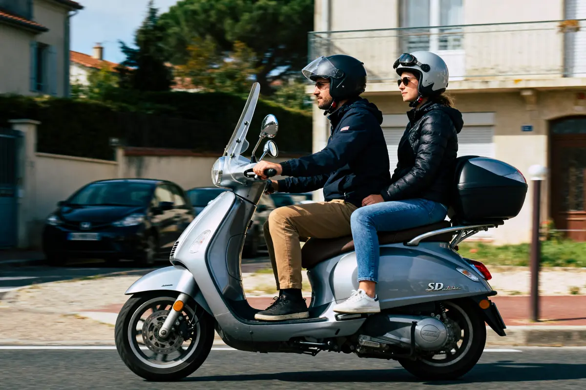 Deux personnes en scooter argenté, portant des casques et des vestes, roulant sur une route urbaine.