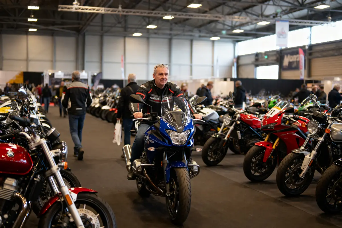 Homme sur moto bleue au salon motocyclettes, entouré de nombreux autres modèles et visiteurs en arrière-plan.