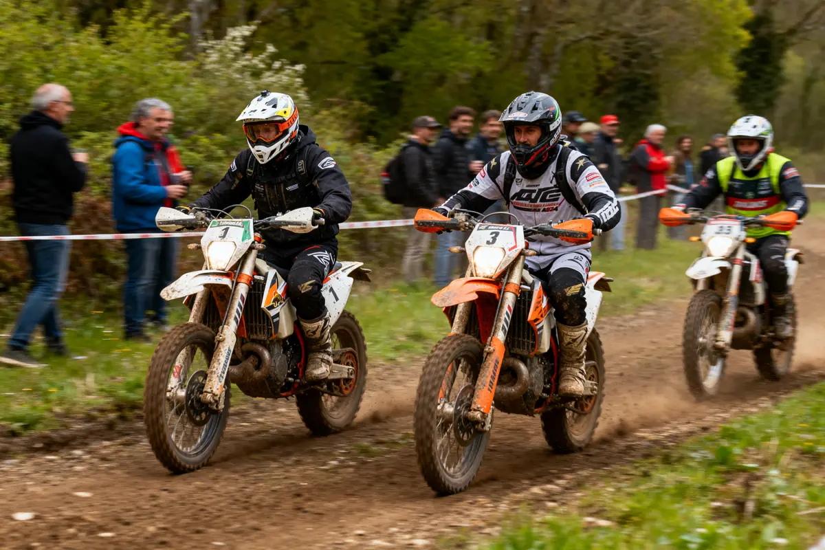 Trois pilotes de motocross en compétition sur un chemin forestier, spectateurs en arrière-plan.