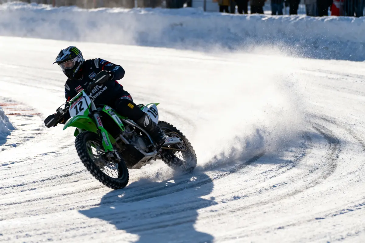 Motard en pleine course sur piste enneigée, créant des gerbes de neige en dérapant.