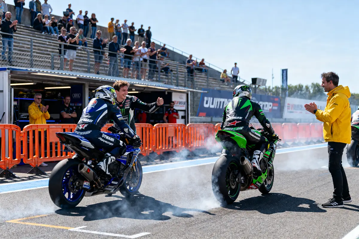 Deux motos font des burnouts, applaudis par des spectateurs et un homme en jaune, lors d'un événement sportif.