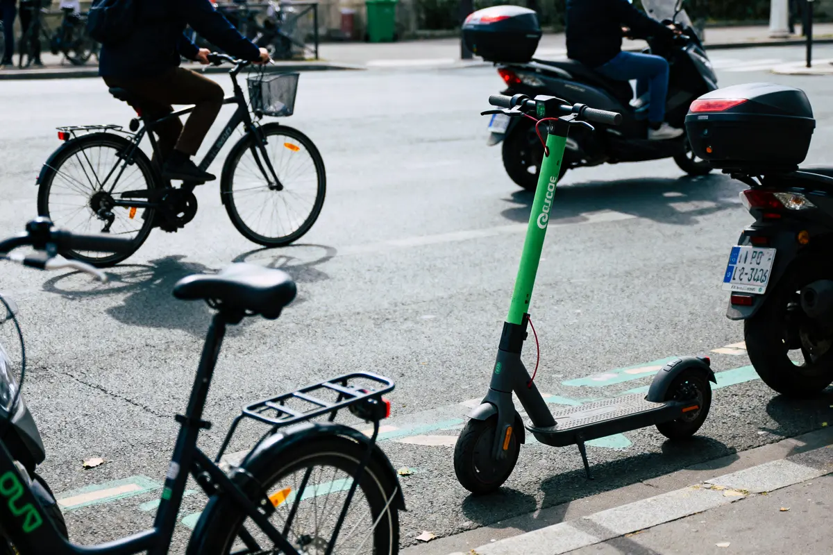 Trottinette verte garée, vélos et scooter circulant sur une route ensoleillée de la ville.