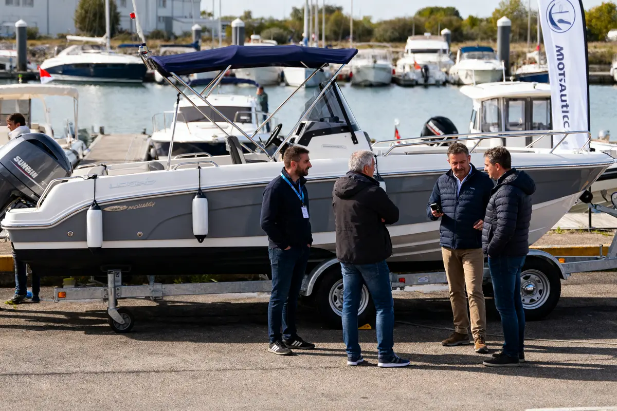 Quatre hommes discutent près d'un bateau gris sur remorque, dans un port avec d'autres bateaux.
