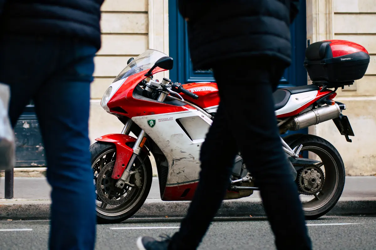 Moto rouge stationnée dans la rue, passants flous au premier plan, mur en pierre en arrière-plan.