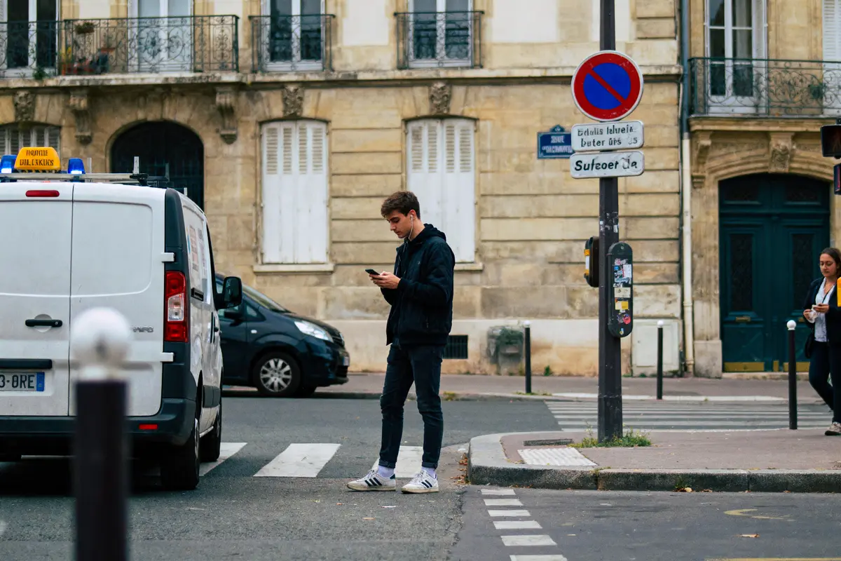 Deux personnes regardent leurs téléphones, debout sur un passage piéton en ville près de véhicules.
