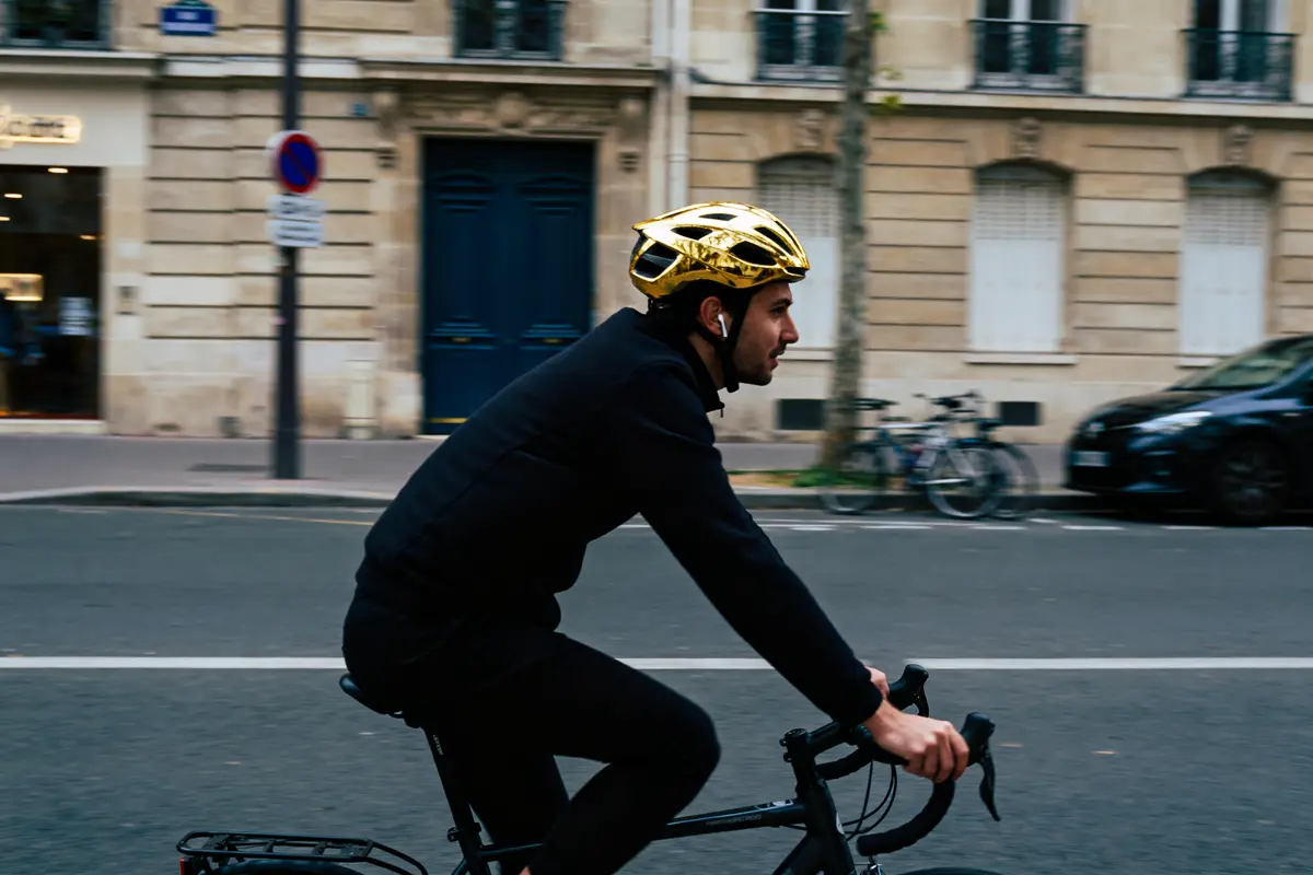 Homme portant casque doré, pédale vélo en ville, bâtiments en arrière-plan.