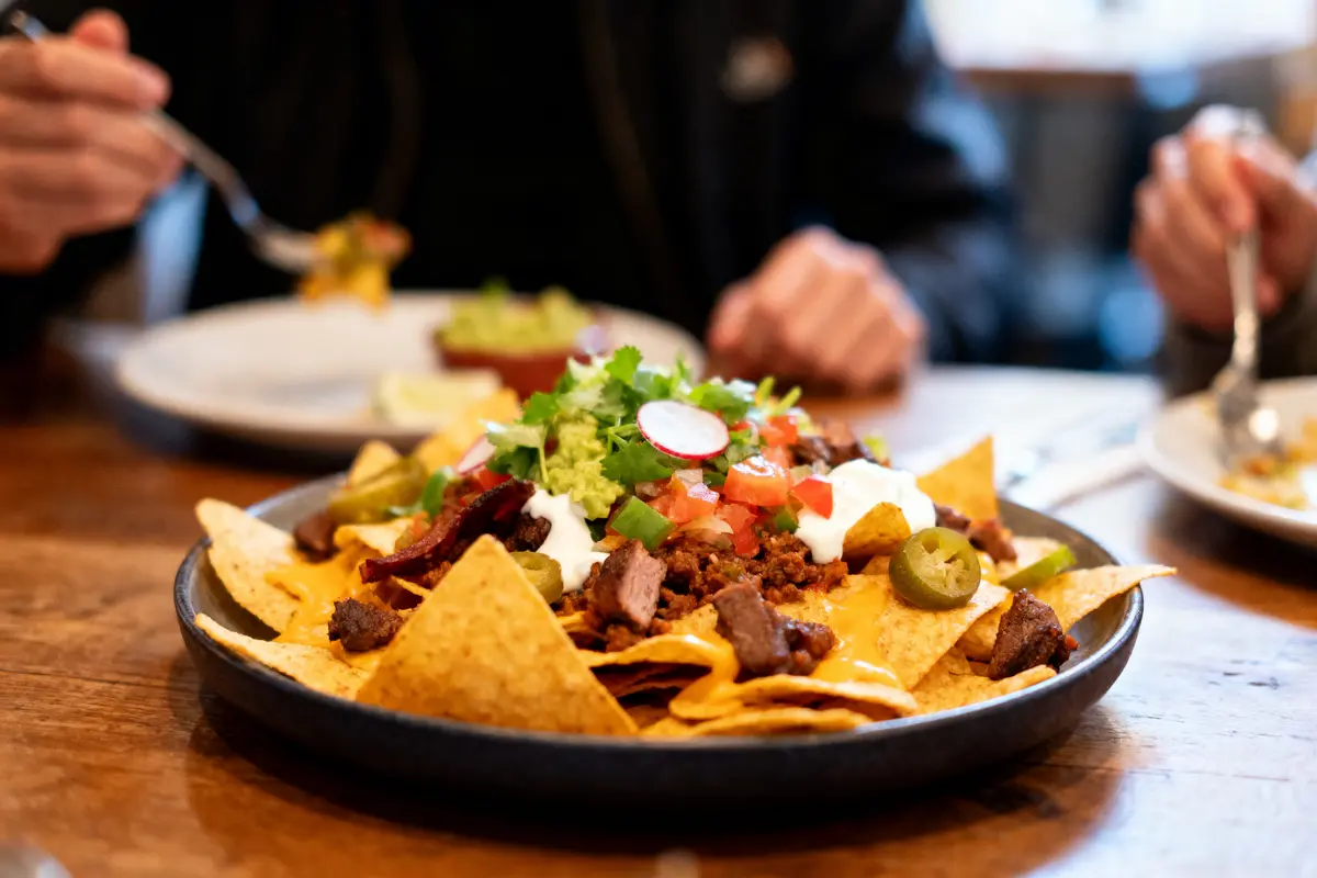 Assiette de nachos avec garnitures variées, servie sur une table en bois. Personnes mangeant en arrière-plan.