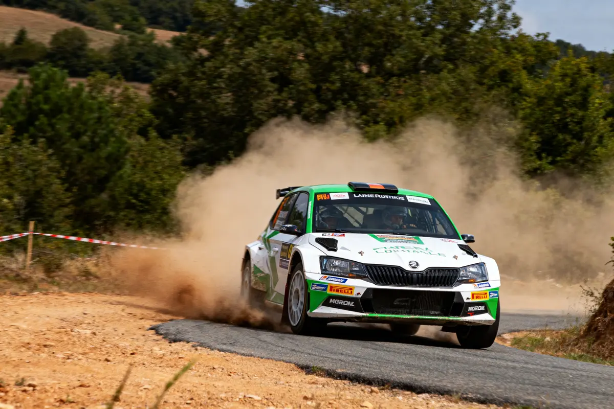 Voiture de rallye blanche sur piste poussiéreuse, entourée de nature et d'arbres en arrière-plan.