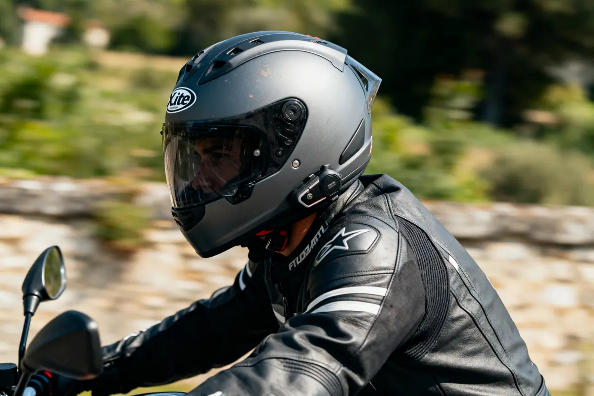 Motard portant un casque gris et une veste en cuir noire, roulant à l'extérieur dans un cadre verdoyant.
