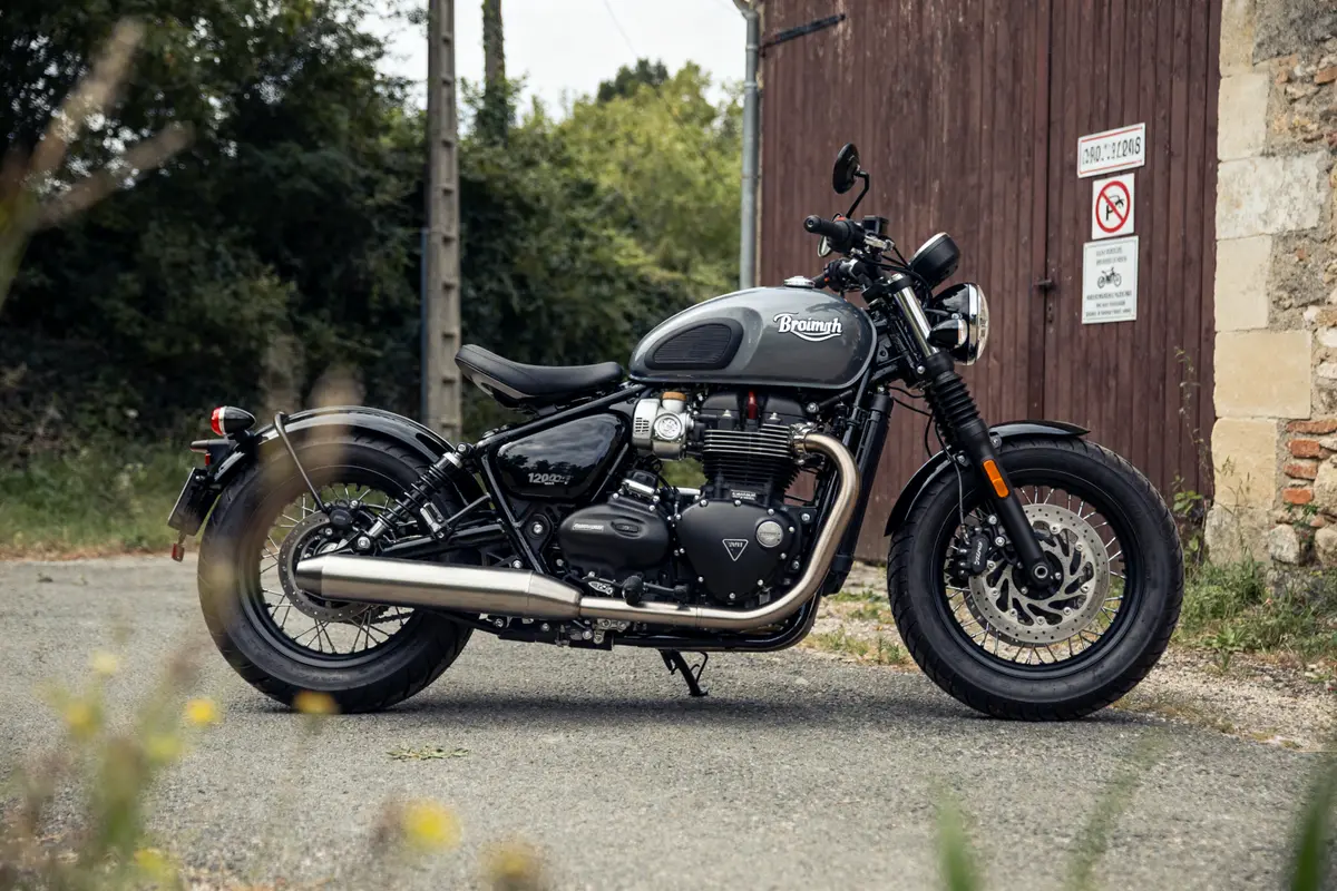Moto Triumph Bonneville Bobber garée devant un bâtiment en bois sur une route rurale