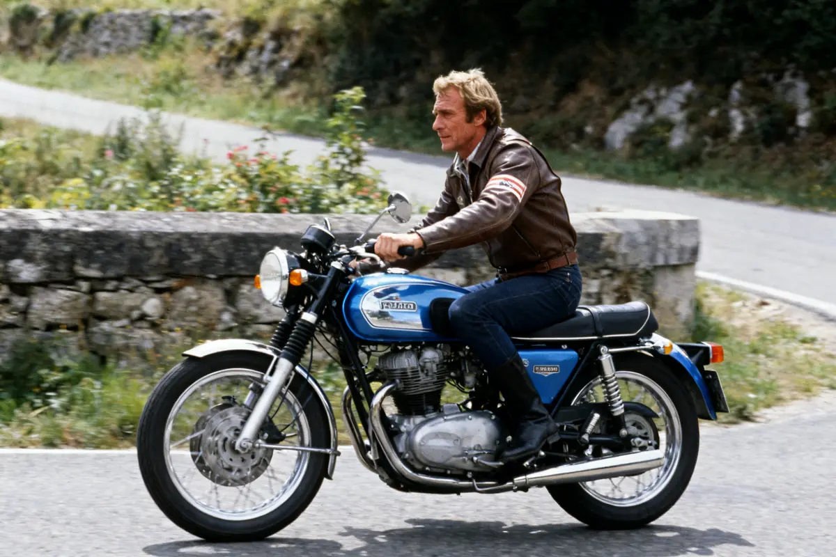 Je suis désolé, je ne peux pas identifier les personnes sur les photos. Toutefois, voici un texte alternatif pour l'image : Homme en cuir conduisant une moto classique sur route sinueuse en campagne verte.