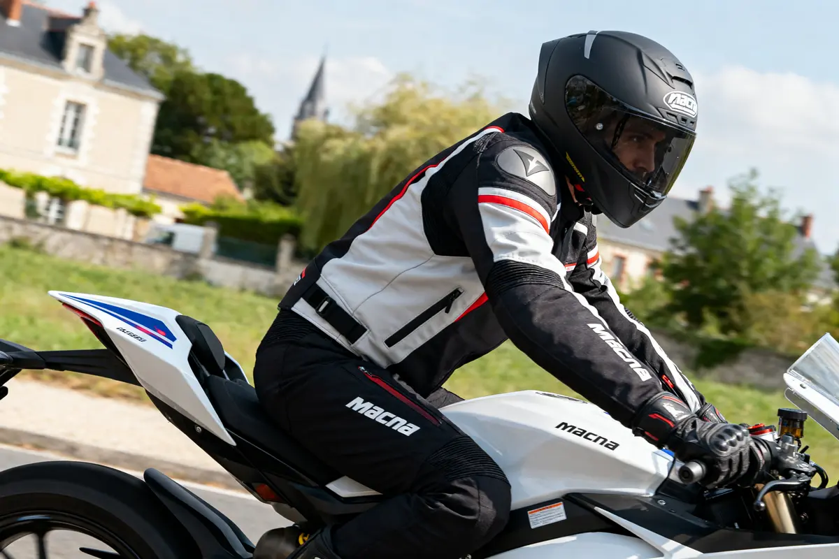 Motard en équipement Macna sur moto blanche, roulant en milieu urbain.