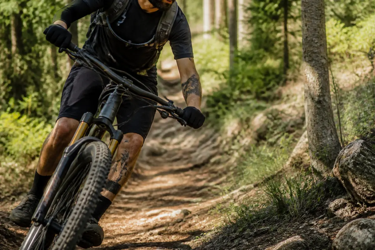 Cycliste en VTT descendant un sentier forestier, vêtu de noir, entouré de verdure et de troncs d'arbres.